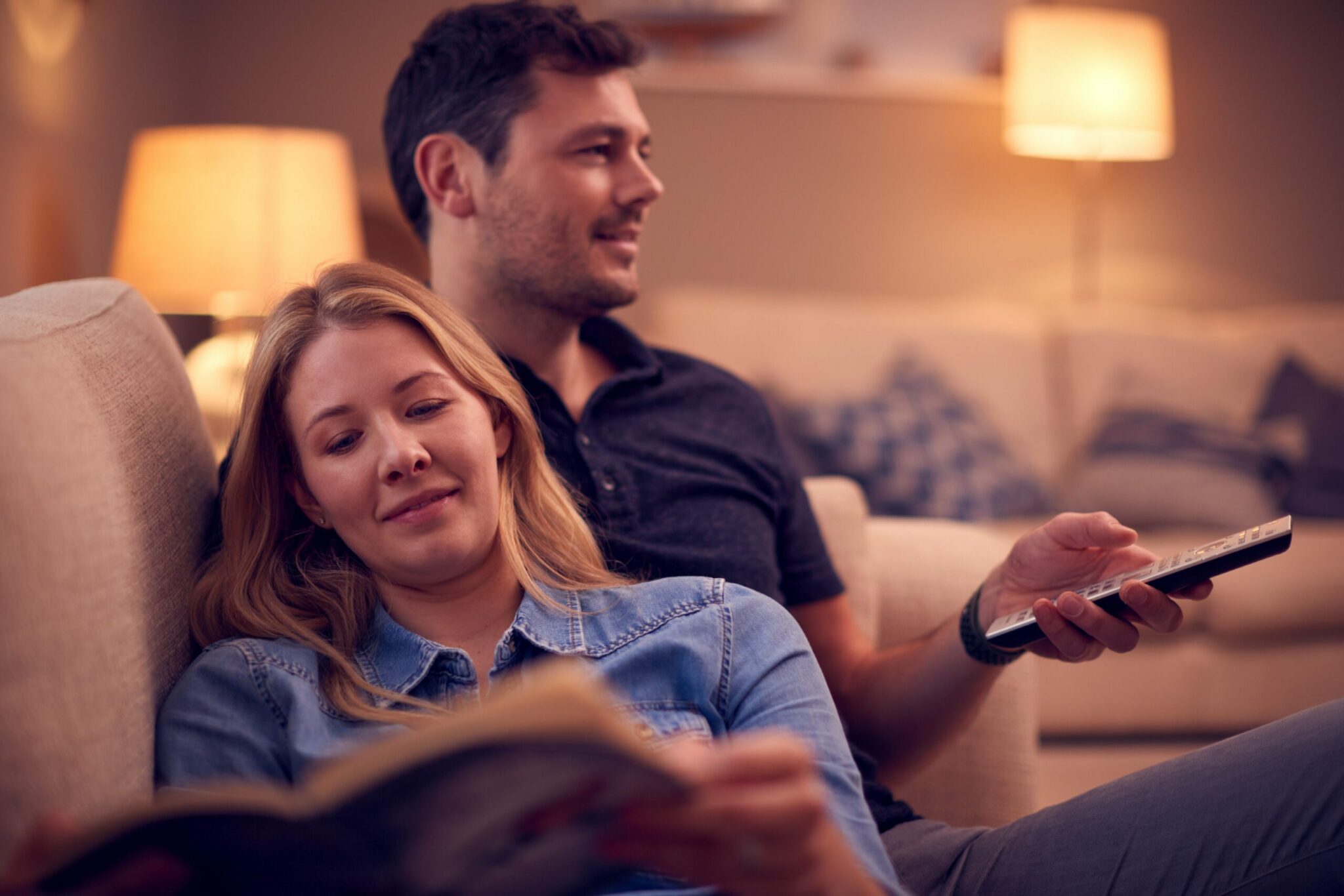 Evening Shot Of Couple Relaxing On Sofa At Home As Woman Reads Book And Man Watches TV E27