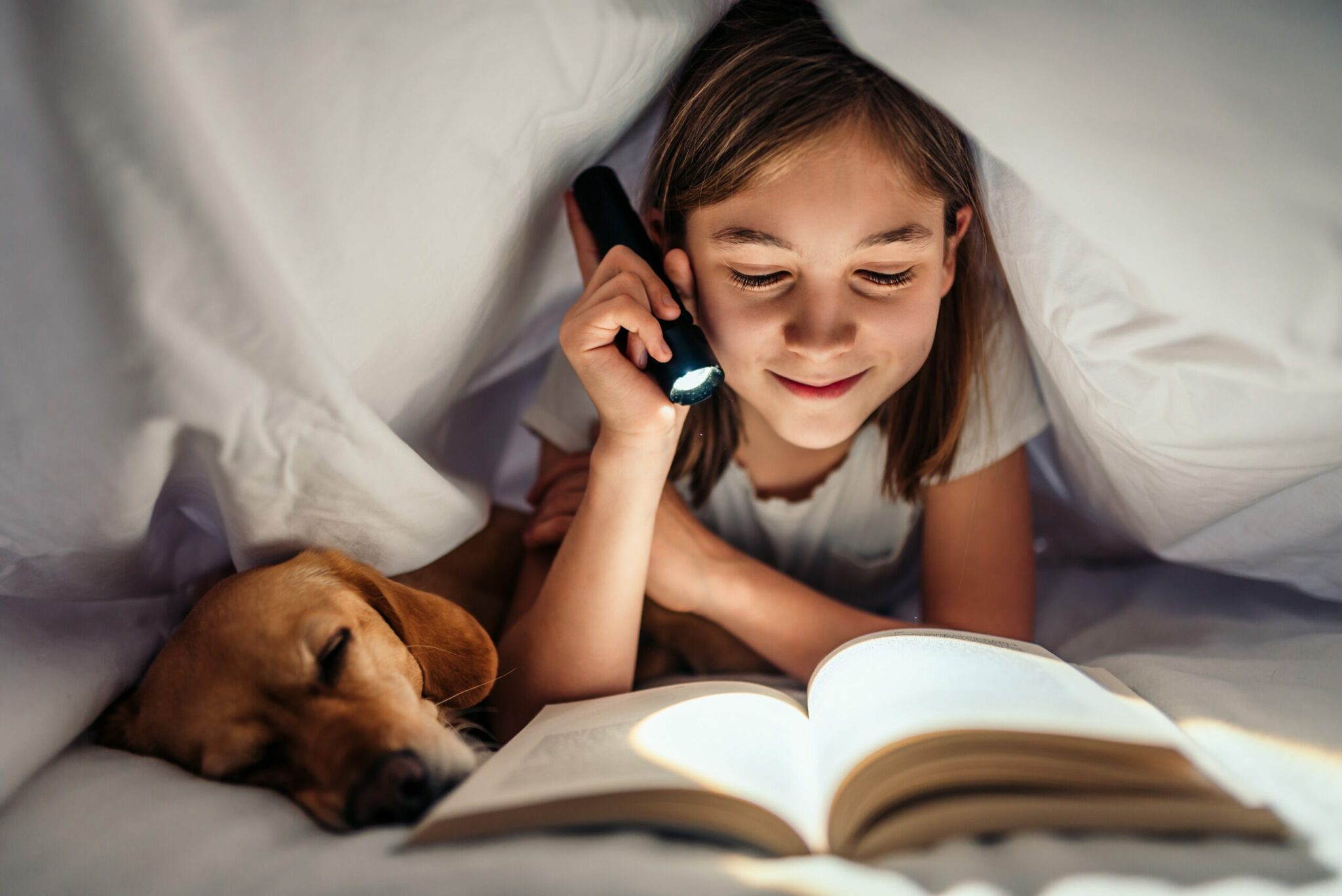 Girl lying in the bed with her dog under blanket reading book late at night Latarka